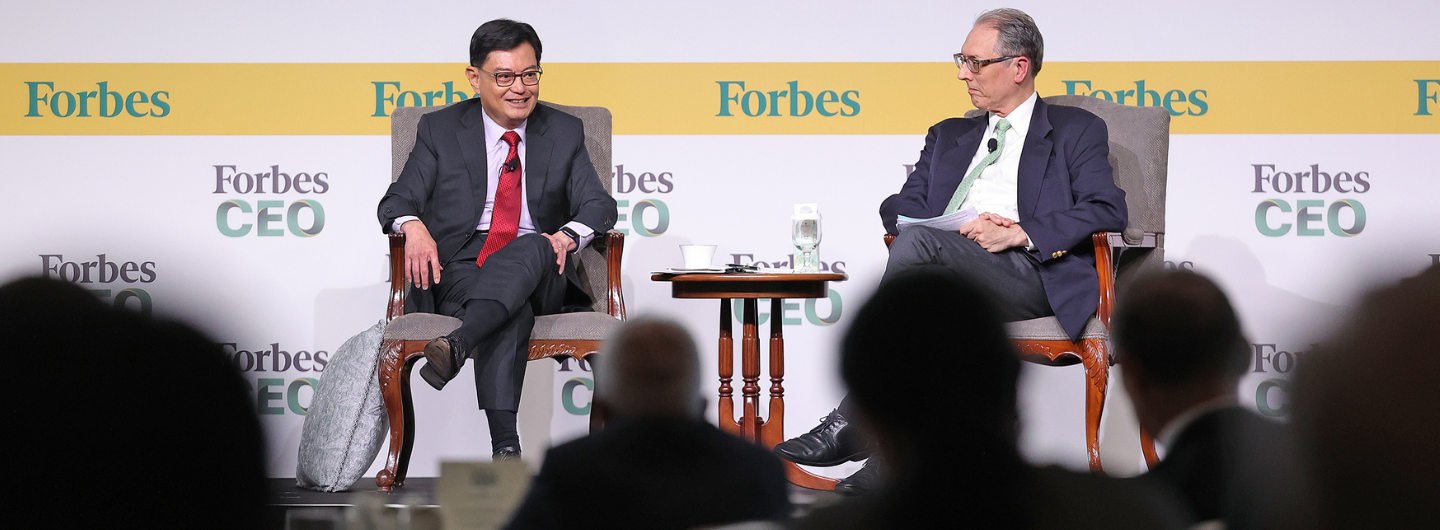 Two men on stage at Forbes CEO event, seated in chairs. Audience is visible.